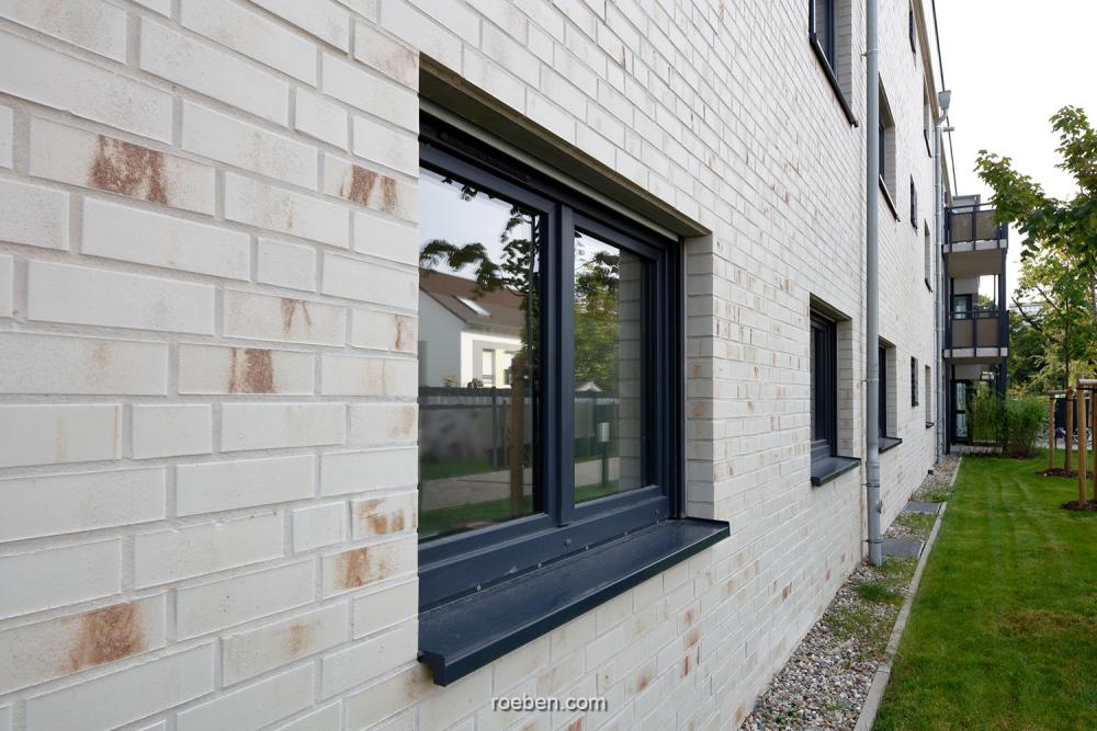 Ziegelwand mit schwarzen Fenstern, daneben ein Rasenstreifen.