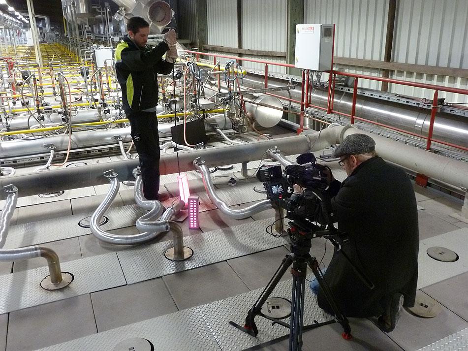 Zwei Männer in einer Fabrikhalle, einer filmt mit einer Kamera auf einem Stativ.