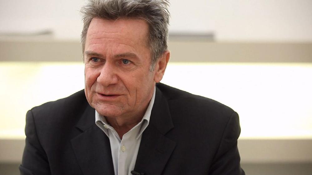 Mann im Anzug spricht, ernster Gesichtsausdruck, helle Kulisse.