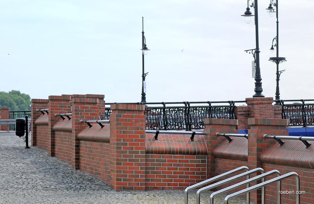 Rote Backsteinmauer an einer Promenade mit Laternen und Geländer.