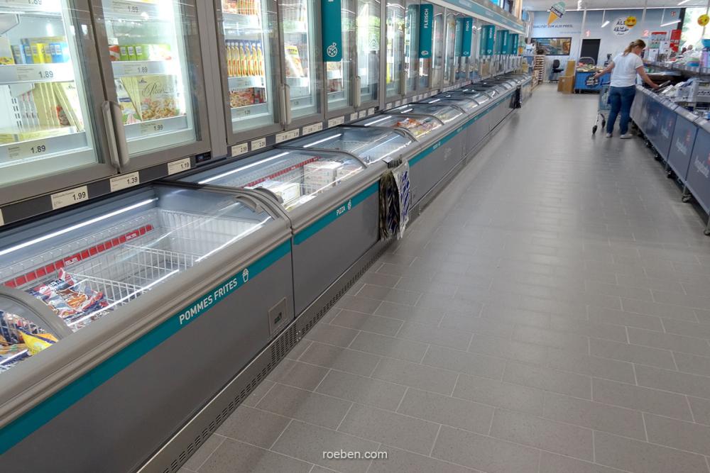 Gänge mit Tiefkühltruhen im Supermarkt, teils gefüllt, Person im Hintergrund.