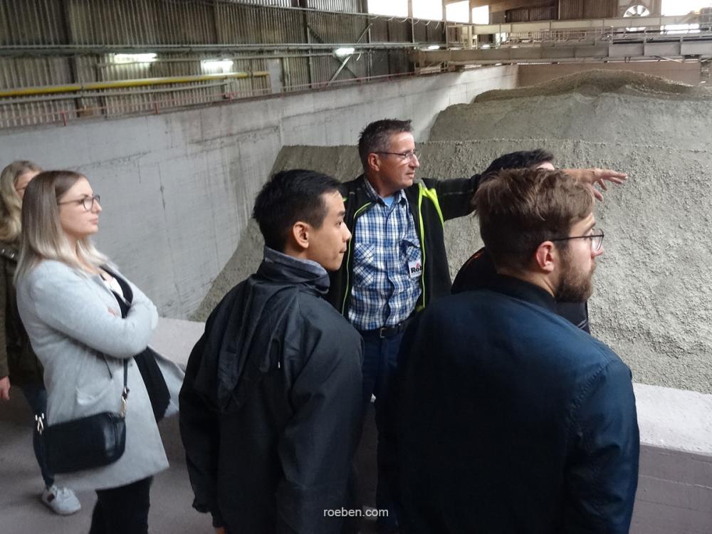 Gruppe von Personen in einer Fabrikhalle, ein Mann zeigt auf etwas.