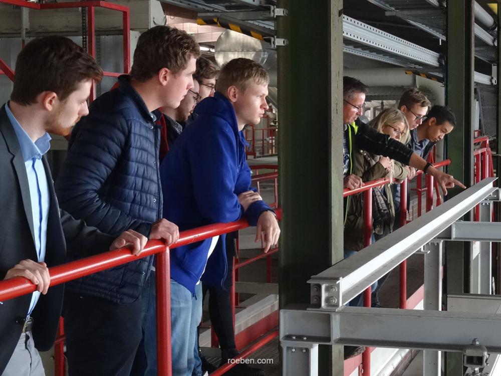 Gruppe von Menschen blickt über ein Geländer in einer industriellen Umgebung.