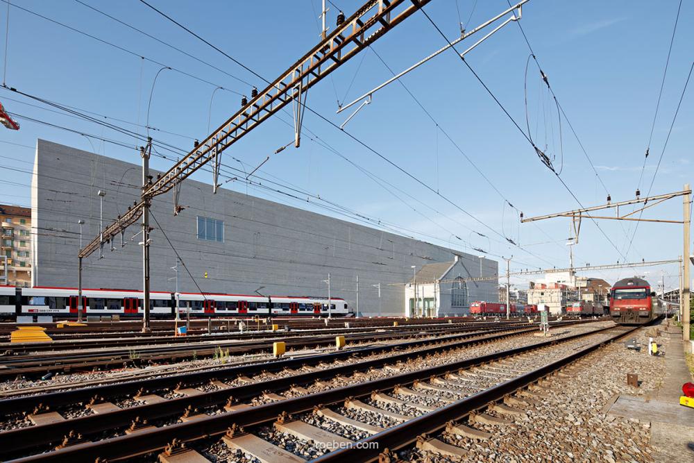 Bahnhof mit roten und weißen Zügen, blauer Himmel, Gleise im Vordergrund.