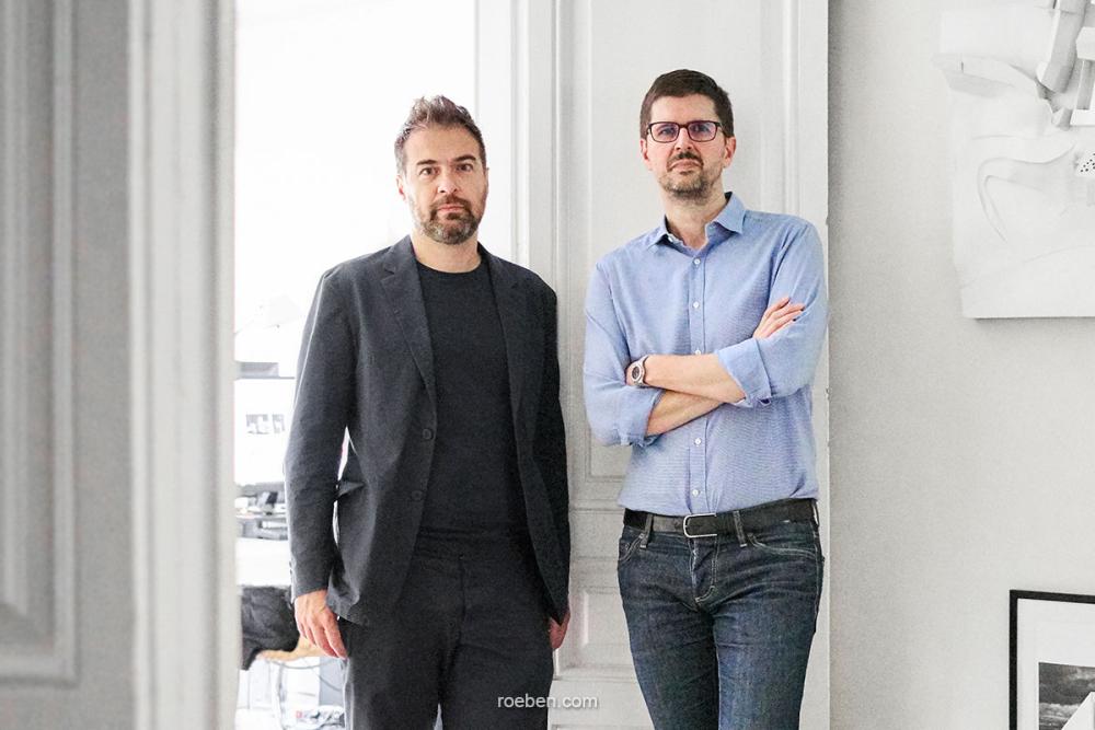 Zwei Männer in einem Büro, einer trägt Anzug, der andere Hemd und Jeans.